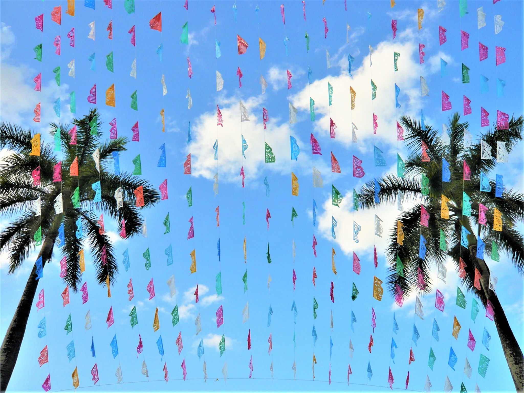 Colourful streamer decorations and two palm trees set against a blue sky at a local Mexican festival.