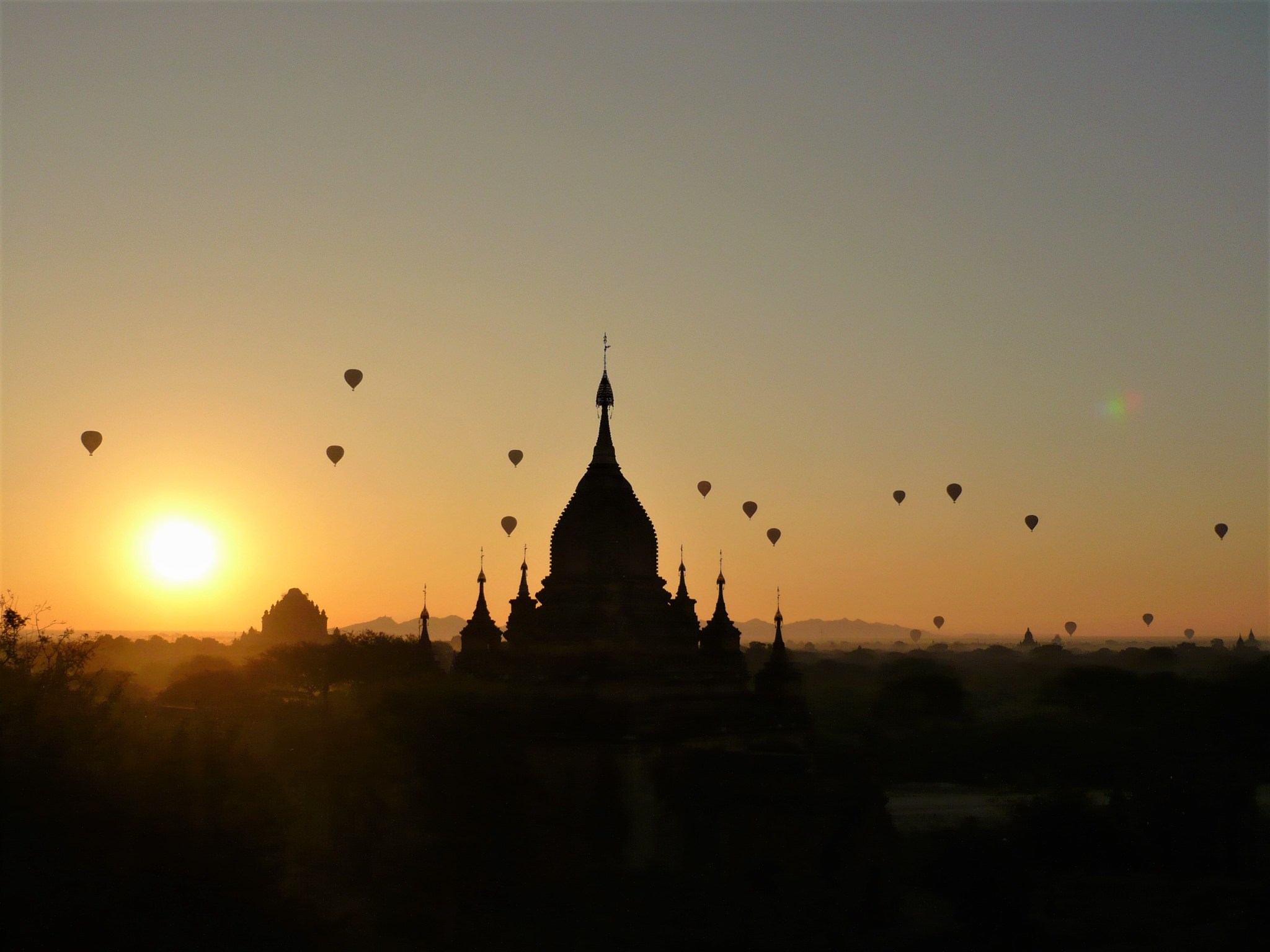 Several hot-air balloons flying at sunrise over the silhouetted temples of Bagan.