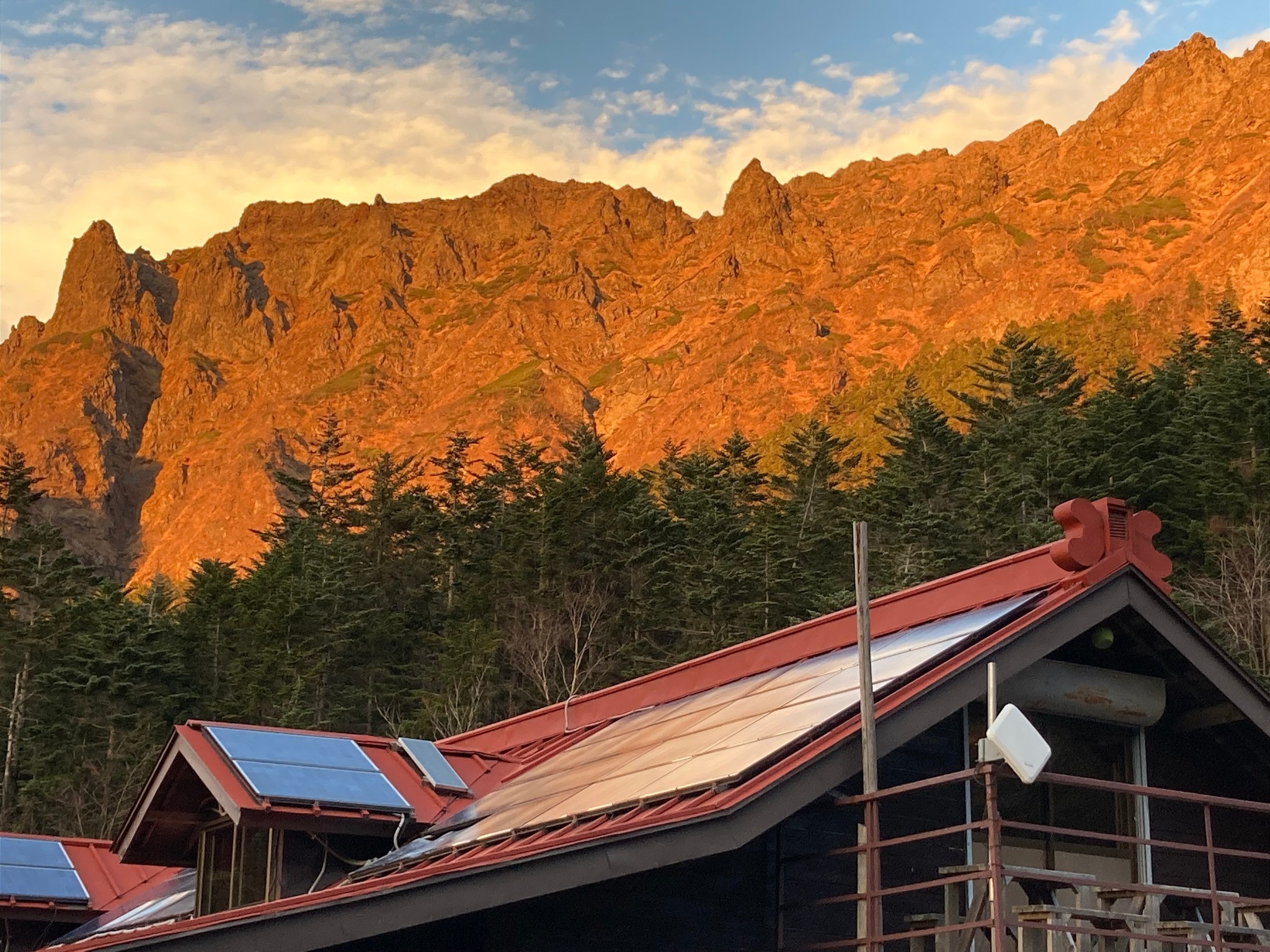 The red-coloured face of Yatsugatake mountain range at sunset, fronted by evergreens and the roof of a mountain lodge.