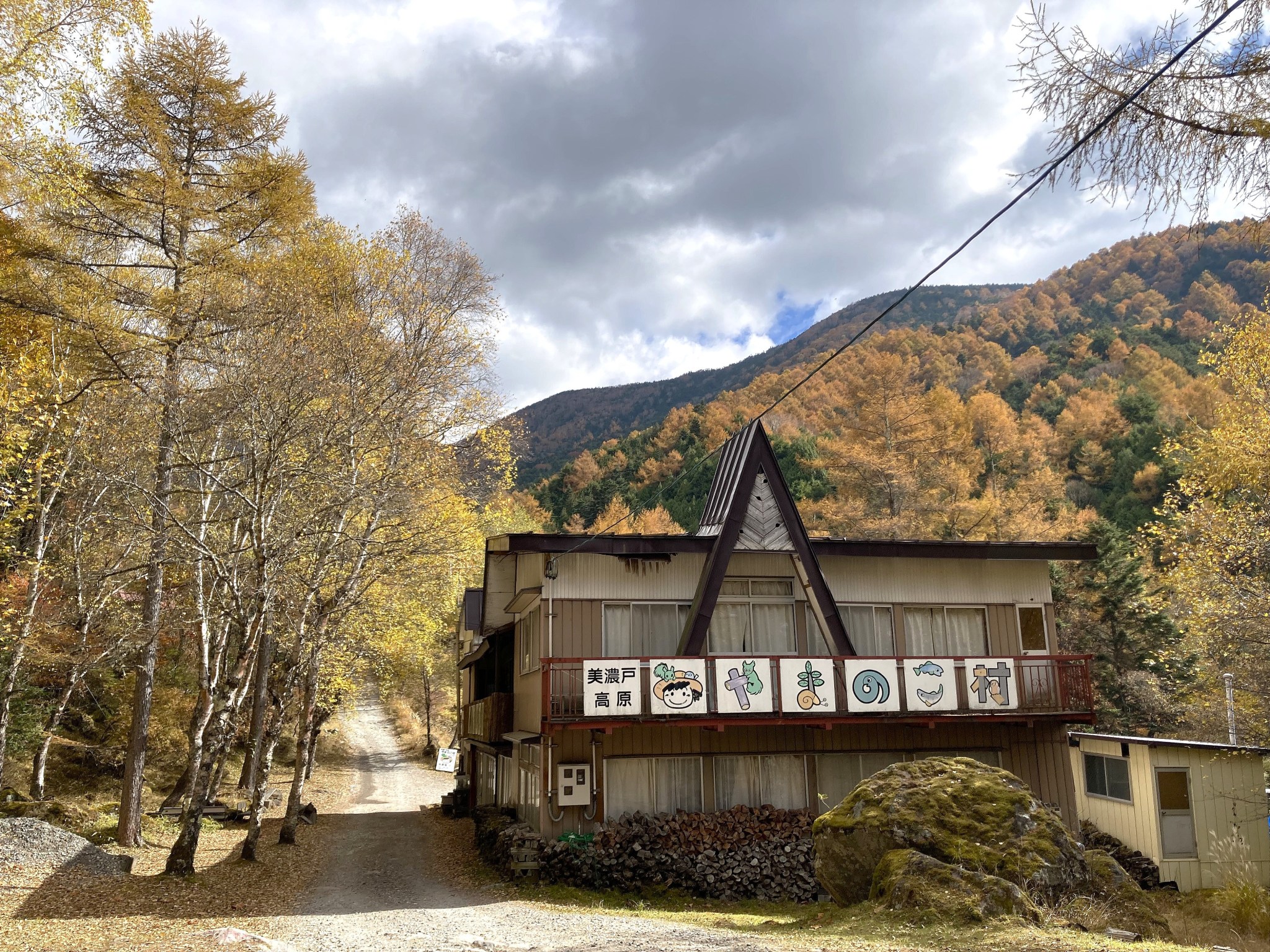A closed-up worn wooden building beside a path leading into an autumnal forest with yellow and orange trees beneath a cloudy sky.