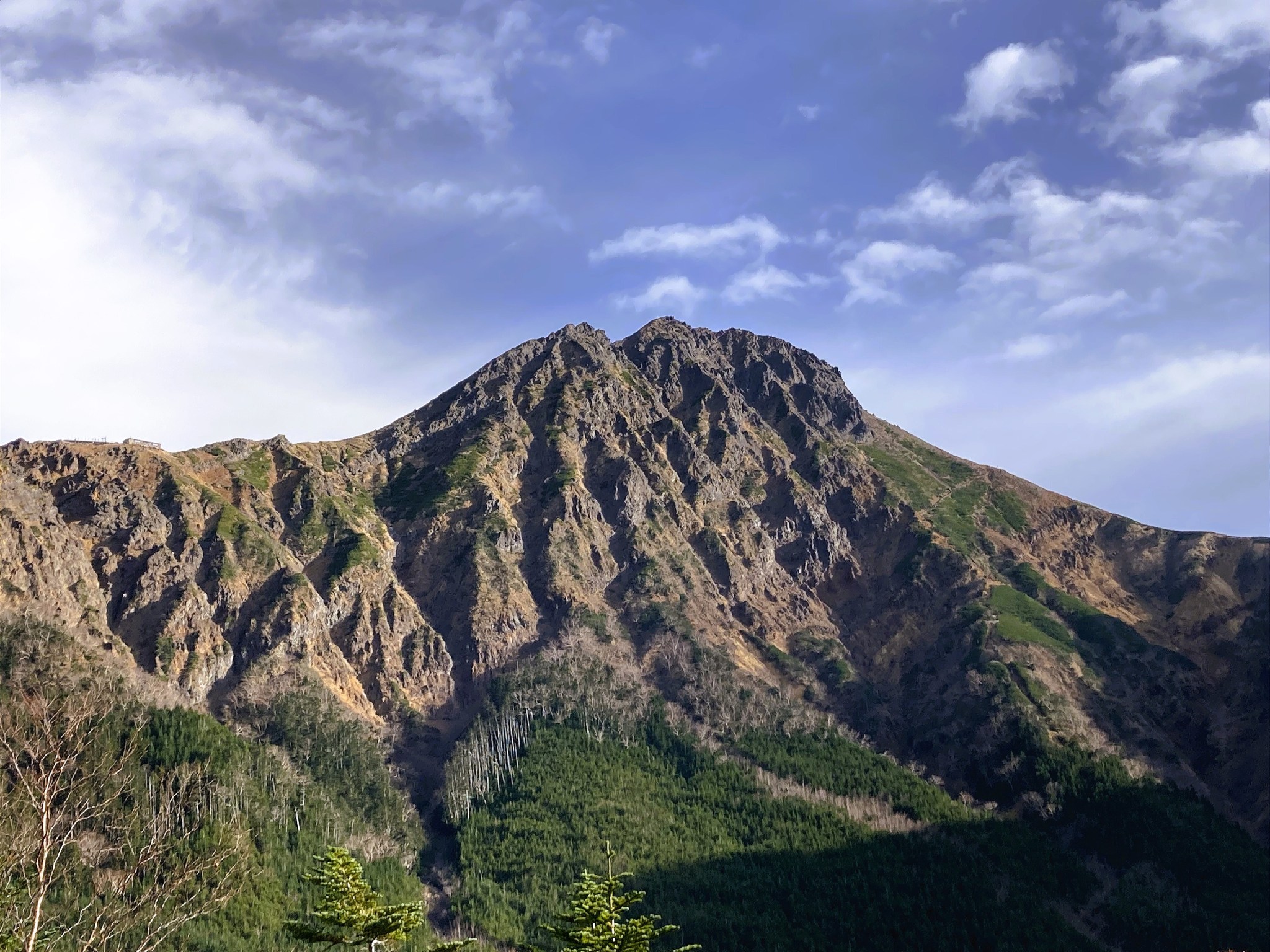 The tan and grey peak of Mount Akadake rising above an evergreen forest, and sat beneath a cool blue sky.