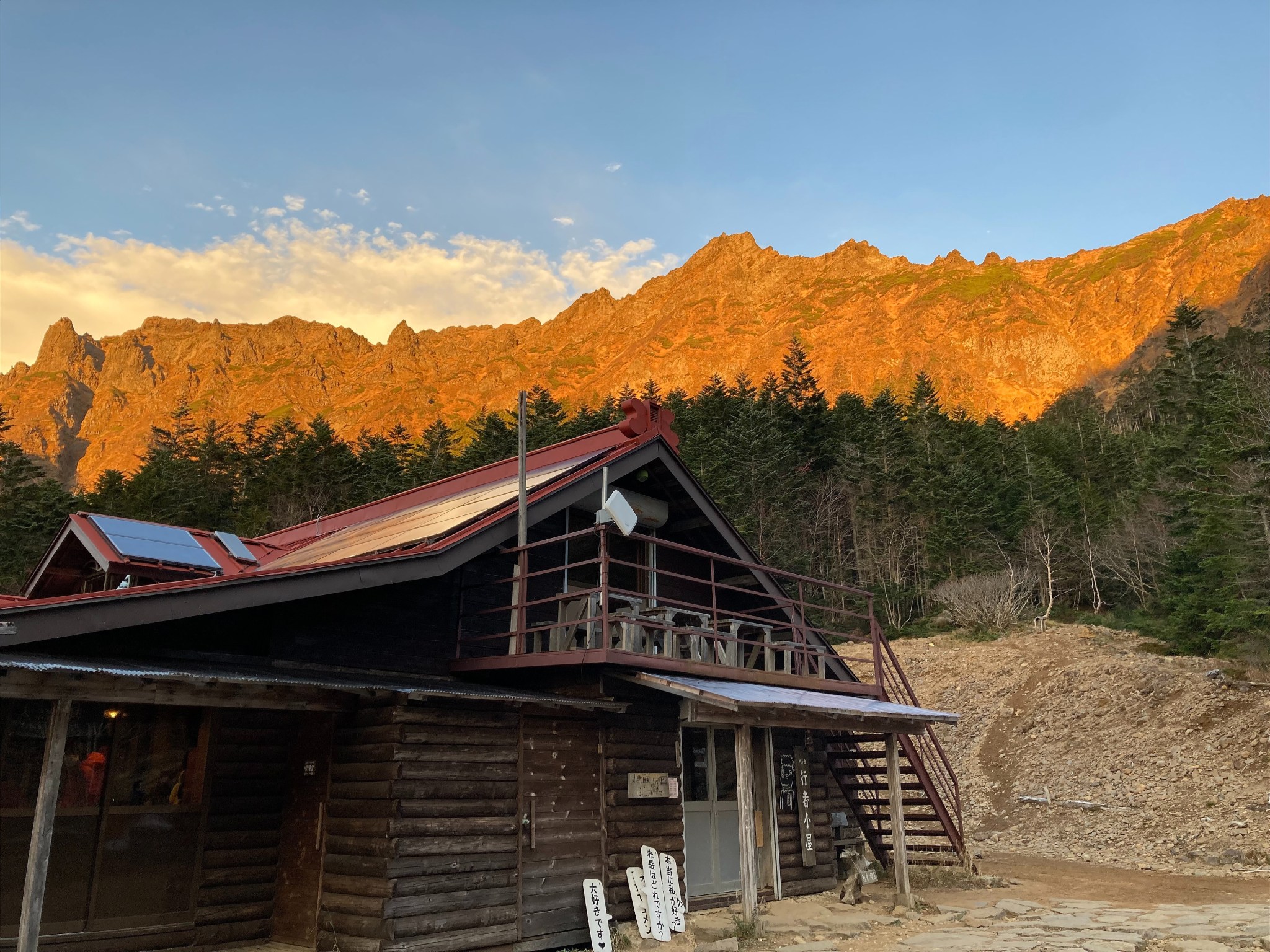 The red-coloured face of Yatsugatake mountain range at sunset, fronted by evergreens and the roof of a mountain lodge.