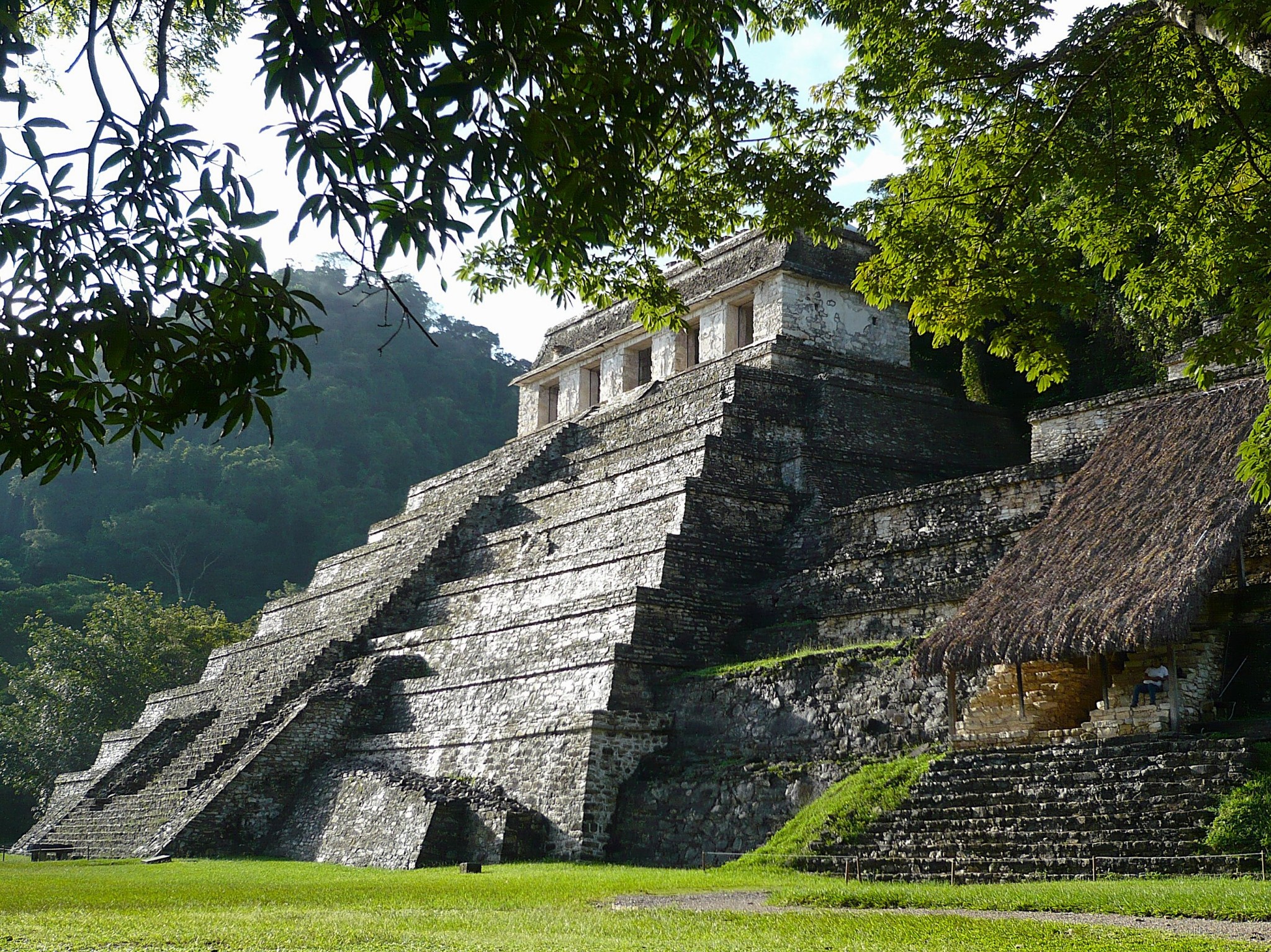 The stone Temple of the Inscriptions surrounded by green grass and trees at the ancient Mayan archaeological site of Palenque in Mexico.