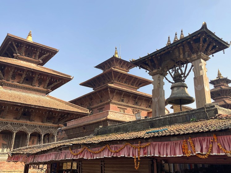 A collection of ancient buildings, spires and a giant bell sitting beneath a pale blue sky in Patan Durbar Square, Nepal.  