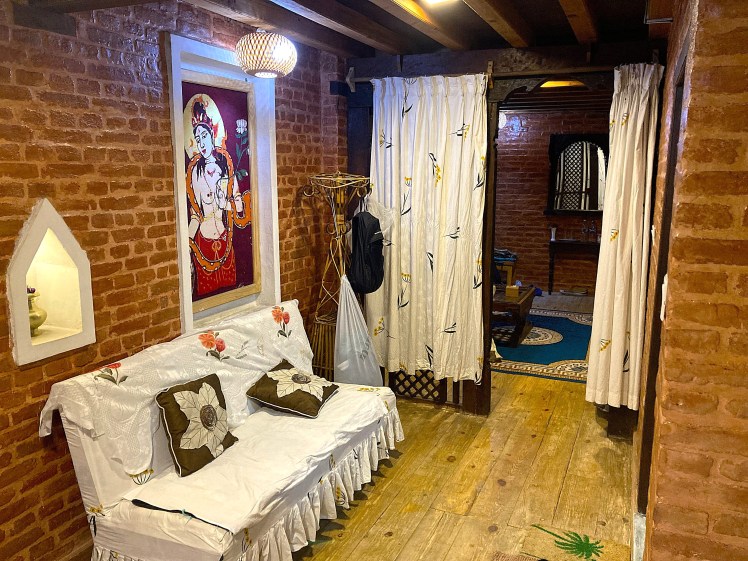 The brick and wood interior of a traditional Nepalese hotel room in Patan containing a white sofa, curtains and a large painting.
