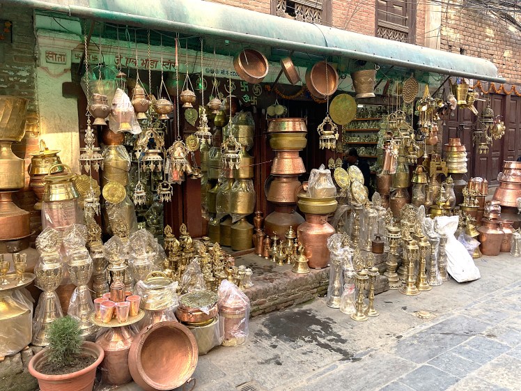 A traditional shopfront in Patan, Nepal with a green awning covering a collection of brass pots, urns and other metal objects.