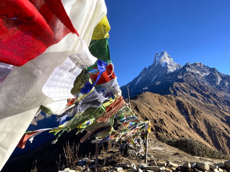 A Himalayan ridgeline adorned with colourful Tibetan prayer flags with Mount Machapuchare in the background highlighted by a bright blue sky.