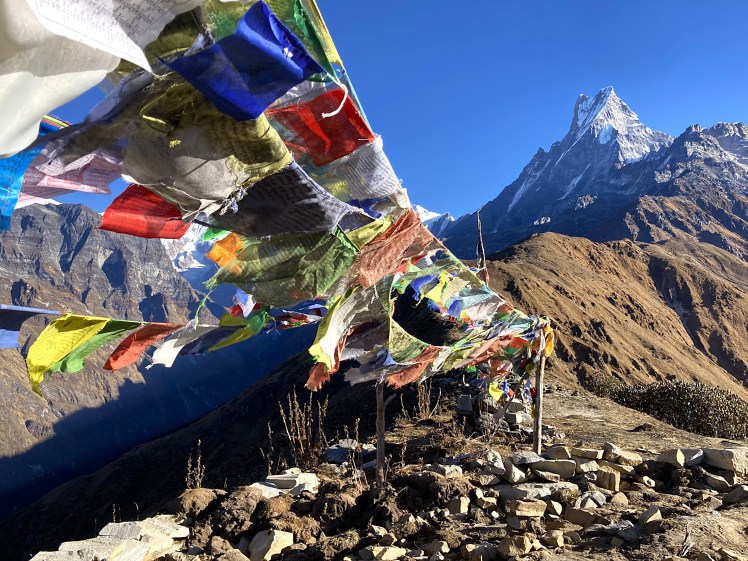 A Himalayan ridgeline adorned with colourful Tibetan prayer flags with Mount Machapuchare in the background highlighted by a bright blue sky.